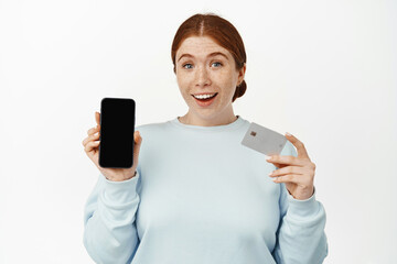 Image of excited smiling ginger girl shows her phone screen, empty mobile display and bank or discount credit card, standing in casual clothing against white background