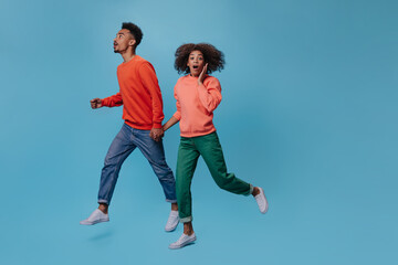 Fototapeta na wymiar Surprised woman and man holding hands and running on blue background. Shocked girl in green pants and man in jeans jump and move on isolated