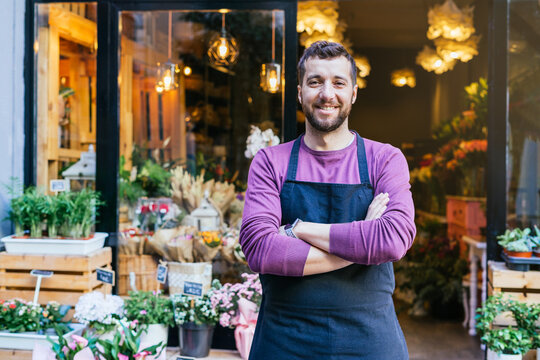 Flower shop owner in an apron has his arms crossed in front of his flower store.