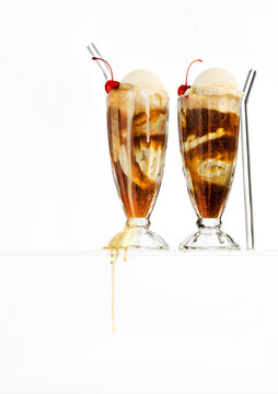 Two Root Beer Floats In Classic Milkshake Glasses Floating On A Glass Surface With Drips. White Background.