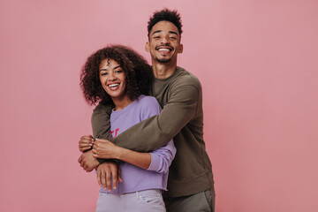 Portrait of happy couple hugging and smiling on pink background. Dark-skinned woman in purple sweater and her boyfriend posing on isolated