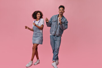 Fototapeta premium Cheerful lady in denim dress and her man dance on pink background. Charming guy in jeans and brunette woman in white tee move on isolated