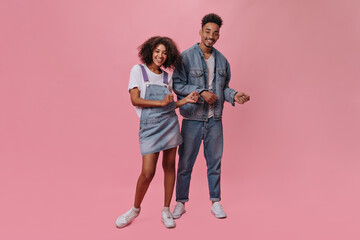 Joyful woman in denim dress and man dance on isolated background. Happy dark-skinned guy and pretty curly girl in blue dress move and smile on pink backdrop