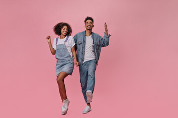 Fototapeta premium Funny gay and girl in jeans outfits dancing on pink background. Cool man and brunette woman in denim jumpsuit move on isolated