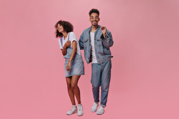 Full length snapshot of man and woman in denim outfits on pink background. Cheerful guy in jeans and cool brunette girl in jumpsuit dance on isolated