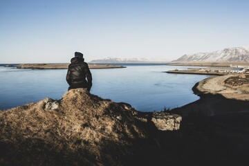 A man in a winter jacket sitting on a rock, watching a winter panorama of snowy Mount Esja ridge and Atlantic ocean. Reykjavik, Iceland. 