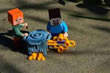 Naklejka premium LEGO Minecraft figures of Steve and Alex opening toy money safe from Kinder Surprise Egg with large yellow key on concrete block in garden. Golden bars are falling out of the bottom.