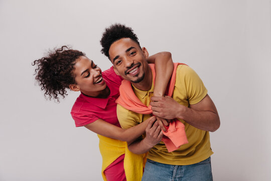 Cool girl and guy hugging and smiling on white background. Close-up porrait of dark-skinned woman in red dress and man in yelllow tee laughing on isolated