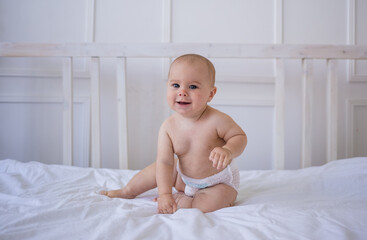 blue-eyed baby girl in a diaper is sitting on a white cotton blanket on the bed in the room