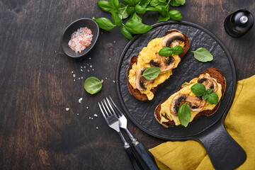 Scrambled eggs with fried mushrooms and basil on bread on black table background. Homemade breakfast or brunch meal - scrambled eggs and mushrooms sandwiches. Top view with copy space