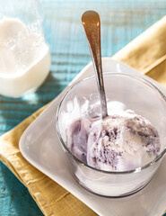 A scoop of berry and vanilla ice cream in a glass dessert dish with a spoon.  Sitting on a white square plate on a yellow linen napkin on a turquoise blue wood surface.  A small glass milk jug at back