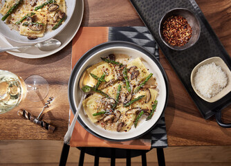 Homemade mushroom ravioli with asparagus in a bowl on a wooden table with a beautiful rust colored linen beneath it. Bowl has red pepper flakes and parmigiana in small bowls next to it. 