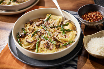Homemade mushroom ravioli with asparagus in a bowl on a wooden table with a beautiful rust colored linen beneath it. Bowl has red pepper flakes and parmigiana in small bowls next to it.  - obrazy, fototapety, plakaty