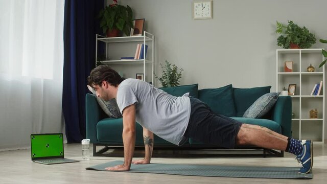 Young man in T-shirt doing push-ups from the floor in living room, using fitness lesson on laptop computer, green screen, chroma key. Remote online sport during coronavirus lockdown isolation at home.