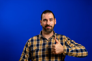 Happy man giving thumbs up sign like hands gesture. Hipster male with beard in blue plaid checkered shirt Isolated on gray studio Background. Negative face expression, human emotion, body language.