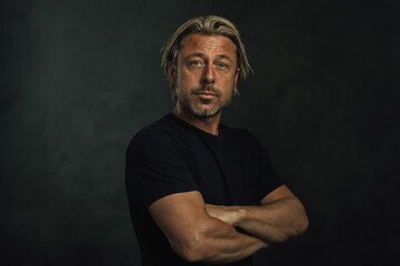 Middle aged tanned blonde man with arms crossed and a stubble in a black t-shirt.