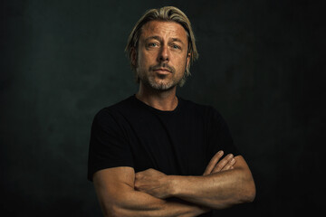 Middle aged tanned blonde man with arms crossed and a stubble in a black t-shirt.