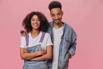 Happy dark skinned man and woman smiling and hugging on pink background. Cool brunette curly girl in white tee and guy in denim outfit laughing on isolated