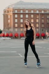 Naklejka premium Full length shot of active smiling woman enjoys extreme sport rides on rollerblades dressed in black comfortable clothes stands outdoors learns rollerskating. People leisure recreation concept