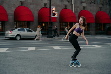 Full length shot of active slim young woman dressed in sportsclothes rides on rollers to strengthen...