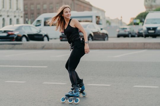Sideways shot of active young woman in good physical shape rollerblades along city streets looks happily behind wears black sportsclothes leads healthy lifestyle. Recreation and hobby concept