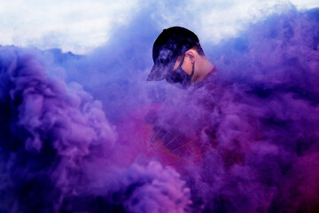 Positive male teenager wit dressed in street style clothes red kimono with chains around neck, black cap and black medical mask . Youth and lifestyle concept. Luxury rap artist in violet smoke.