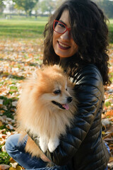 Portrait of a happy woman and her pomeranian pet dog sitting on a ground full of autumn leaves.