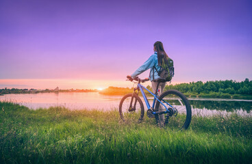 Obraz premium Woman with bicycle near the river at sunset in spring. Landscape with sporty girl with backpack riding a mountain bike, dirt road, green grass, water, purple sky in summer. Sport and travel. Cycle