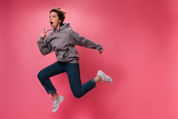 Woman in grey hoodie and jeans jumps on pink background. Emotional teen girl in sweatshirt and denim pants moves on isolated