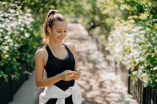 Sport as style of life. Horizontal shot of healthy slim sporty woman focused at smartphone checks information and chooses song from playlist dressed in sportwear poses outdoor breathes fresh air