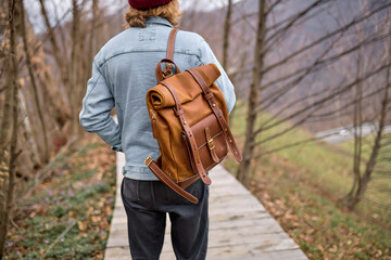 Young man exploring nature landscapes in spring, walking across alone with brown backapack. Handsome Hiker traveling in forests. Mountains in the background, rear view, focus on leather backpack - obrazy, fototapety, plakaty