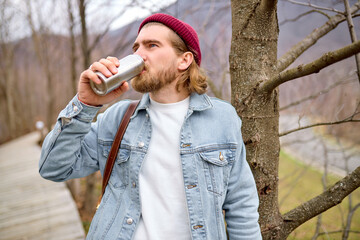 young handsome brutal bearded man traveling in the wild nature with thermos, autumn or spring...