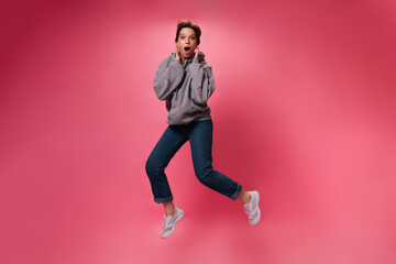 Fototapeta na wymiar Surprised woman in grey hoodie jumping on pink background. Shocked girl in denim pants and sweatshirt moves on isolated backdrop