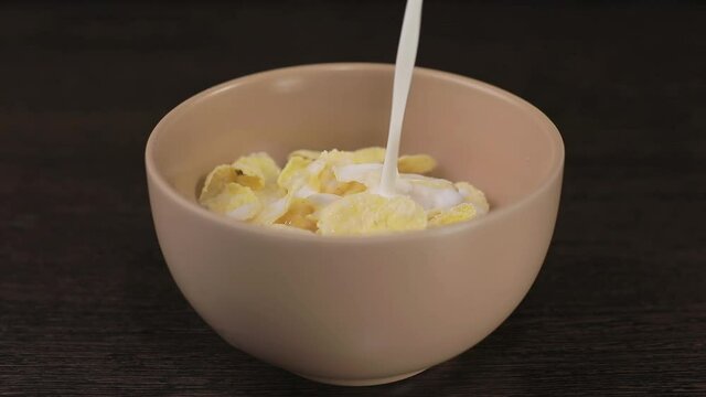 There is a bowl of cornflakes on the table against a black background. Milk begins to pour into it from above. Milk is poured to the very top until the bowl is full.