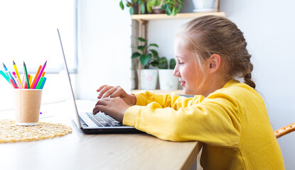 Caucasian blonde school girl, typing on a laptop keyboard while sitting by the window at home, blogging, chatting, viewing social media pages or playing computer games. E-learning, back to school.