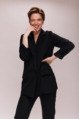 Pretty lady in black outfit winks on isolated background. Charming young woman in dark jacket and pants laughing on white backdrop