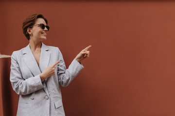 Smiling lady in grey outfit points to place for text. Happy young woman in sunglasses and oversize...