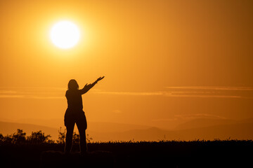 Obraz na płótnie Canvas silhouette of a woman in a field with her arms outstretched as if offering something with a large sun and mountains in the background