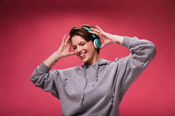 Pretty woman in hoodie listening to music in headphones on pink background. Teen girl in grey...