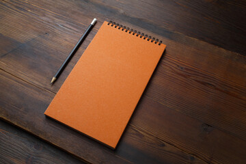 Blank orange notebook and pencil on wood table background. Responsive design mockup.