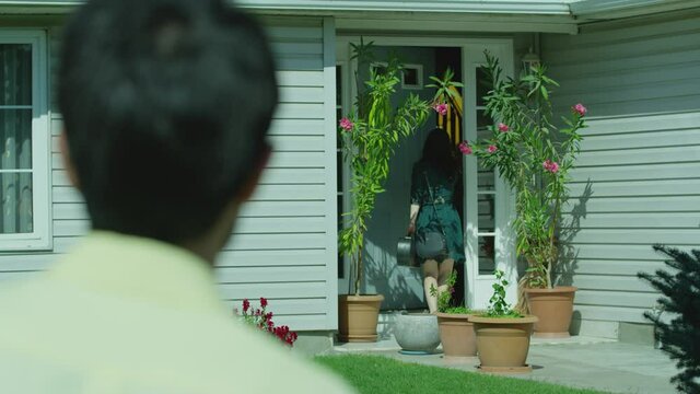 Back rear view stranger man following young girl while she entering home . Musician girl on green dress and violin opens door and enters inside house  boy secretly follows , spying behind her . 

