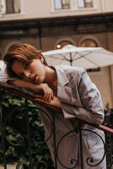 Pretty woman in suit looks into camera and leans on railing. Attractive romantic short-haired girl in jacket and pants thoughtfully poses outside