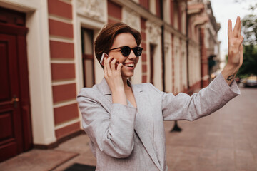 Charming woman in grey outfit talks on phone and waving hand. Happy cheerful girl in oversize jacket and sunglasses smiles and greets outside