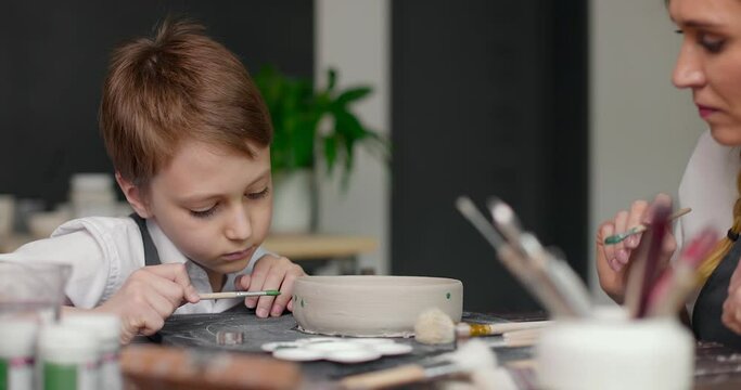 Mom and her son draw a clay bowl together in a pottery studio, workshop. Woman potter teaches the boy to paint on earthenware. Slow motion