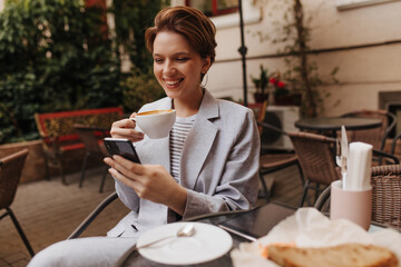 Woman chatting on smartphone and drinking coffee. Pretty lady in grey jacket and tee widely...