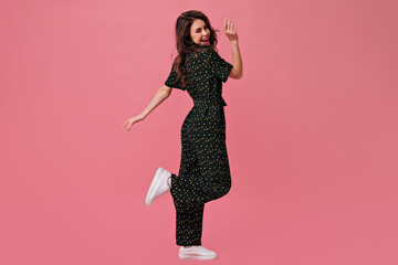 Full length portrait of curly woman in black jumpsuit. Joyful girl with lovely smile poses for camera in black clothes and white sneakers