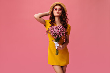 Lady in yellow dress holds bouquet and blows kiss. Cute girl with curly hair in modern clothes with flowers in her hands posing on pink background