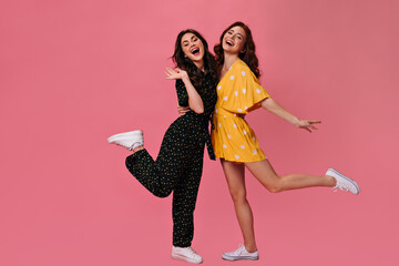 Two Girls in polka dot dresses happily pose on pink background. Cute woman in black overalls hugs beautiful friend in bright yellow dress