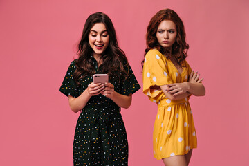 Ginger woman envies her brunette friend chatting on smartphone. Beautiful curly girls in...