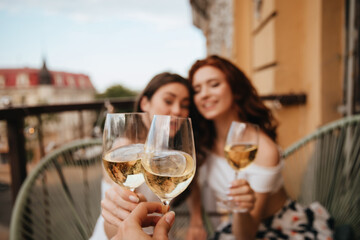 Photo of girls clinking glasses with white wine. Young curly girls on terrace enjoying champagne and posing on balcony